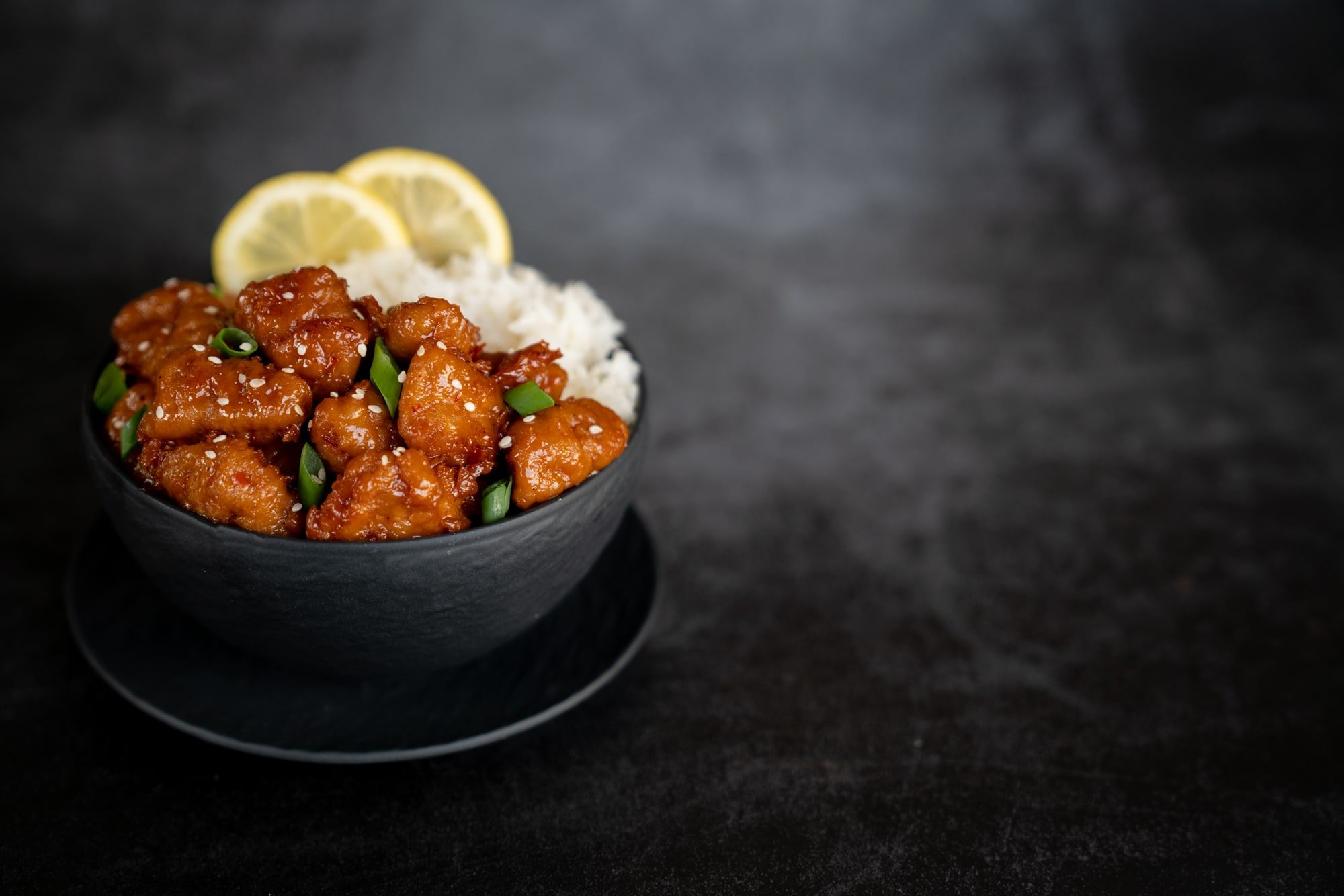 A bowl of glazed plant-based chick'n over rice with lemon and scallion on a dark slate table